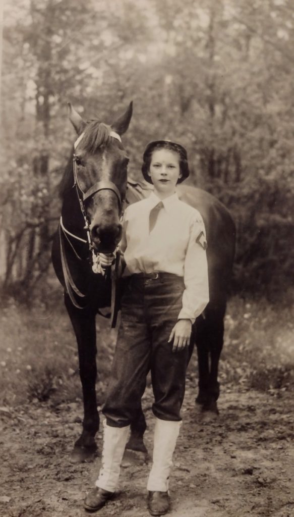 A fifteen year old girl standing next to a horse, holding its bridle; she is wearing a white button-down shirt and tucked tie, riding pants, and gaiters; on her shoulder is a patch representing the head of a horse