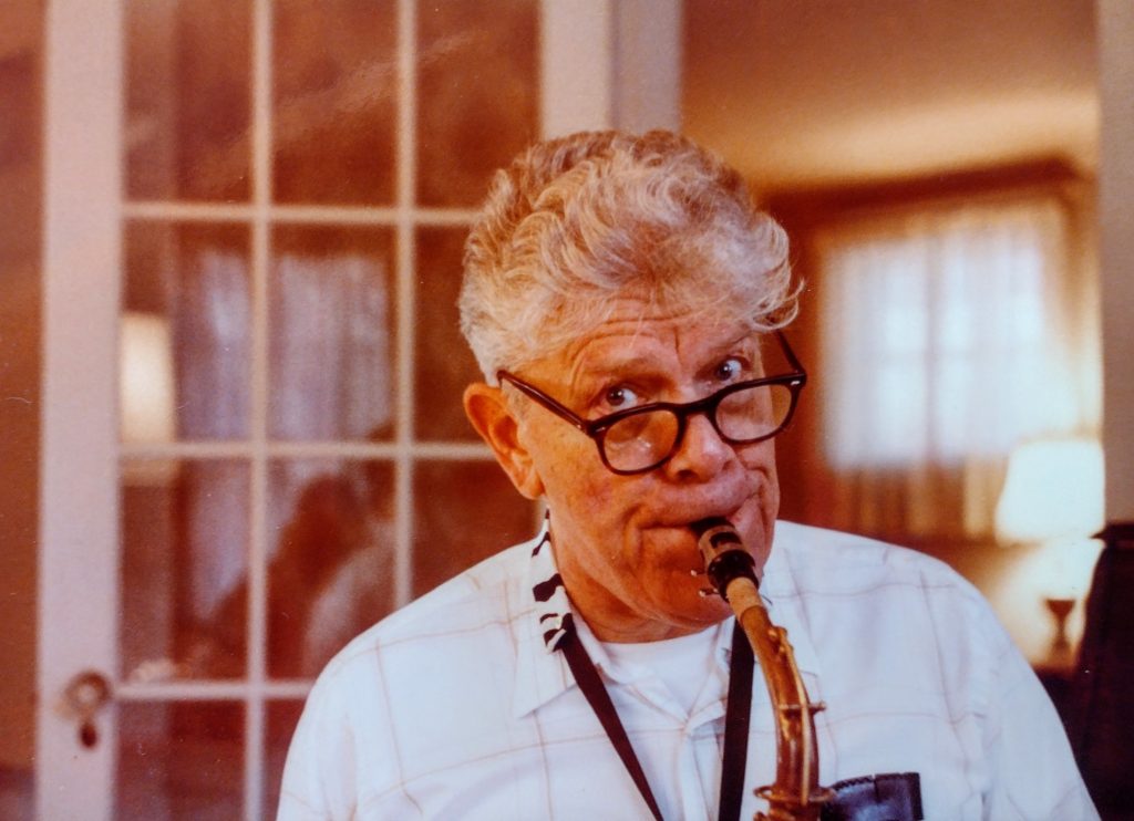 A color photograph of Alton Hall, 64 years old, playing a saxophone in the dining room of a house, looking up at the photographer