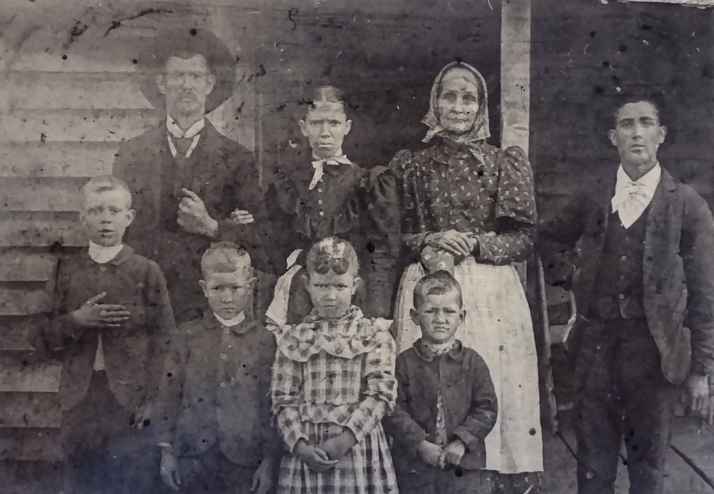 Very old damaged black and white photograph depicting a family of eight, adults and children, standing in front of a wooden shack